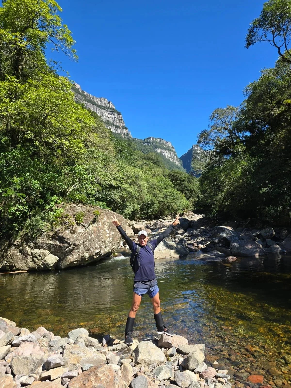 Pessoa feliz com roupas de caminhada em um rio cercado por árvores e rochas, com montanhas ao fundo sob um céu azul claro.
