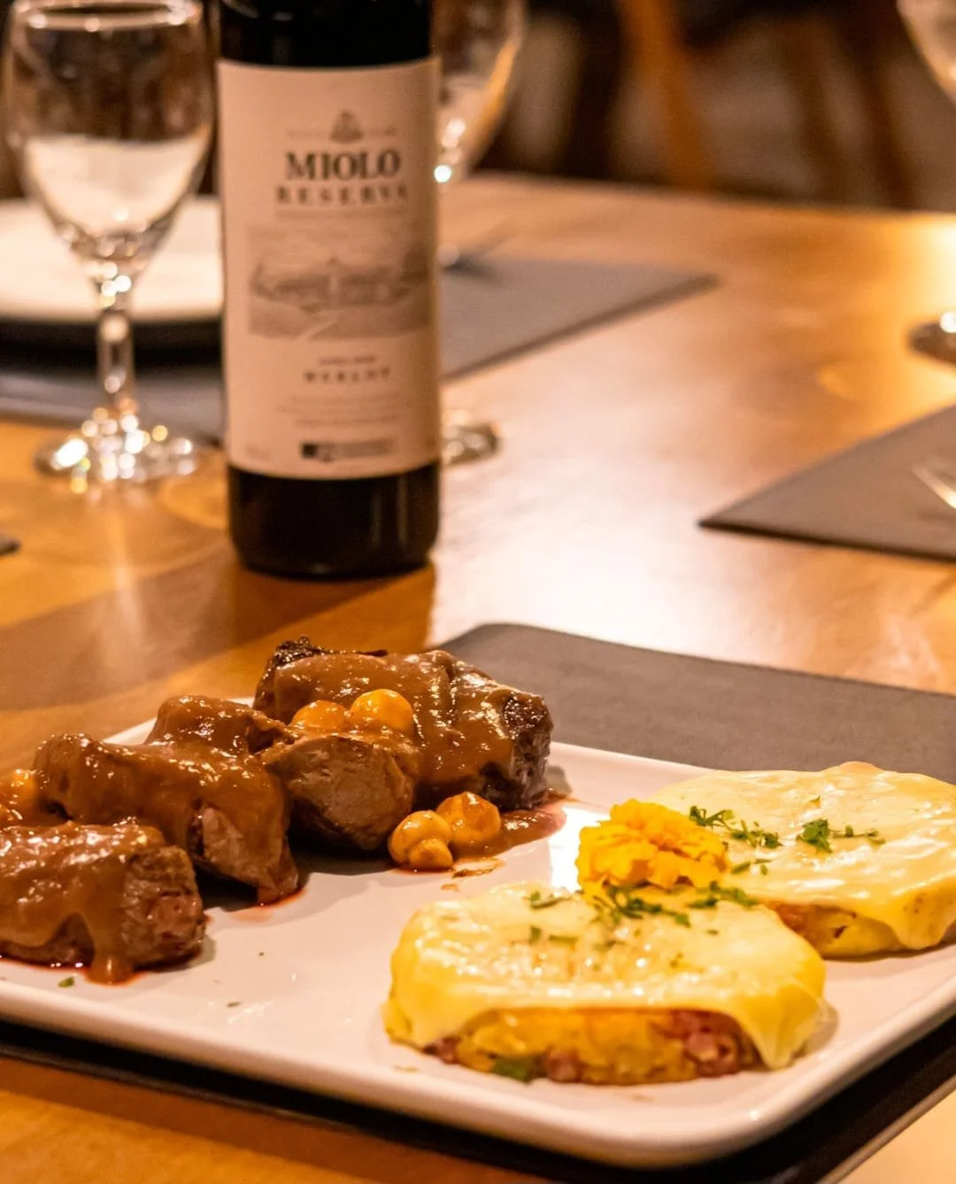 Prato com carne com molho, milho, e dois pedaços de lasanha ou torta com queijo, decorados com salsa, em um jantar com taças de vinho e garrafa de vinho ao fundo.