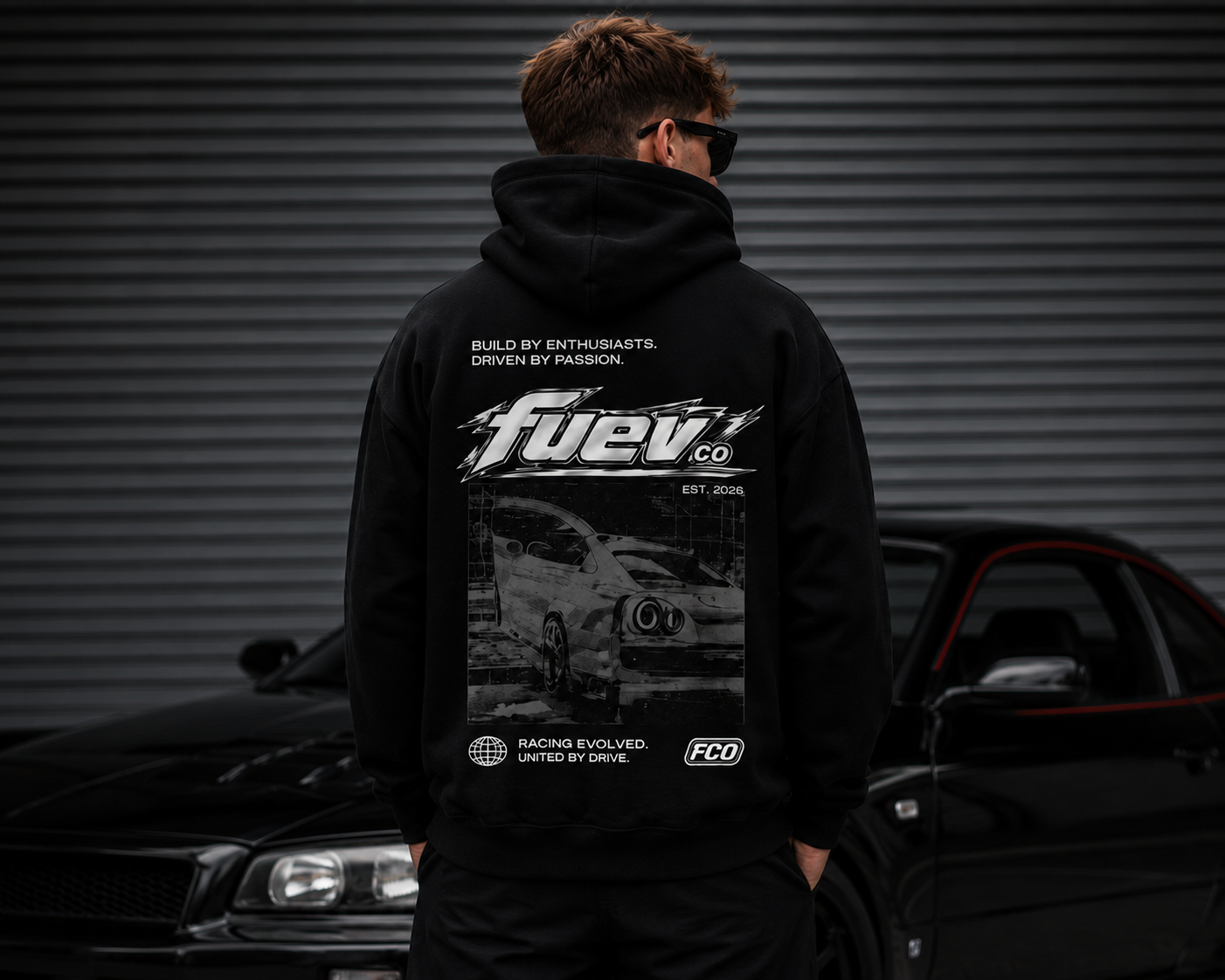 Person wearing a black hoodie with car graphics and text, standing in front of a black sports car and corrugated metal background.