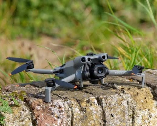 A gray quadcopter drone with a camera mounted on the front, resting on a mossy brick surface outdoors amidst green grass and vegetation.
