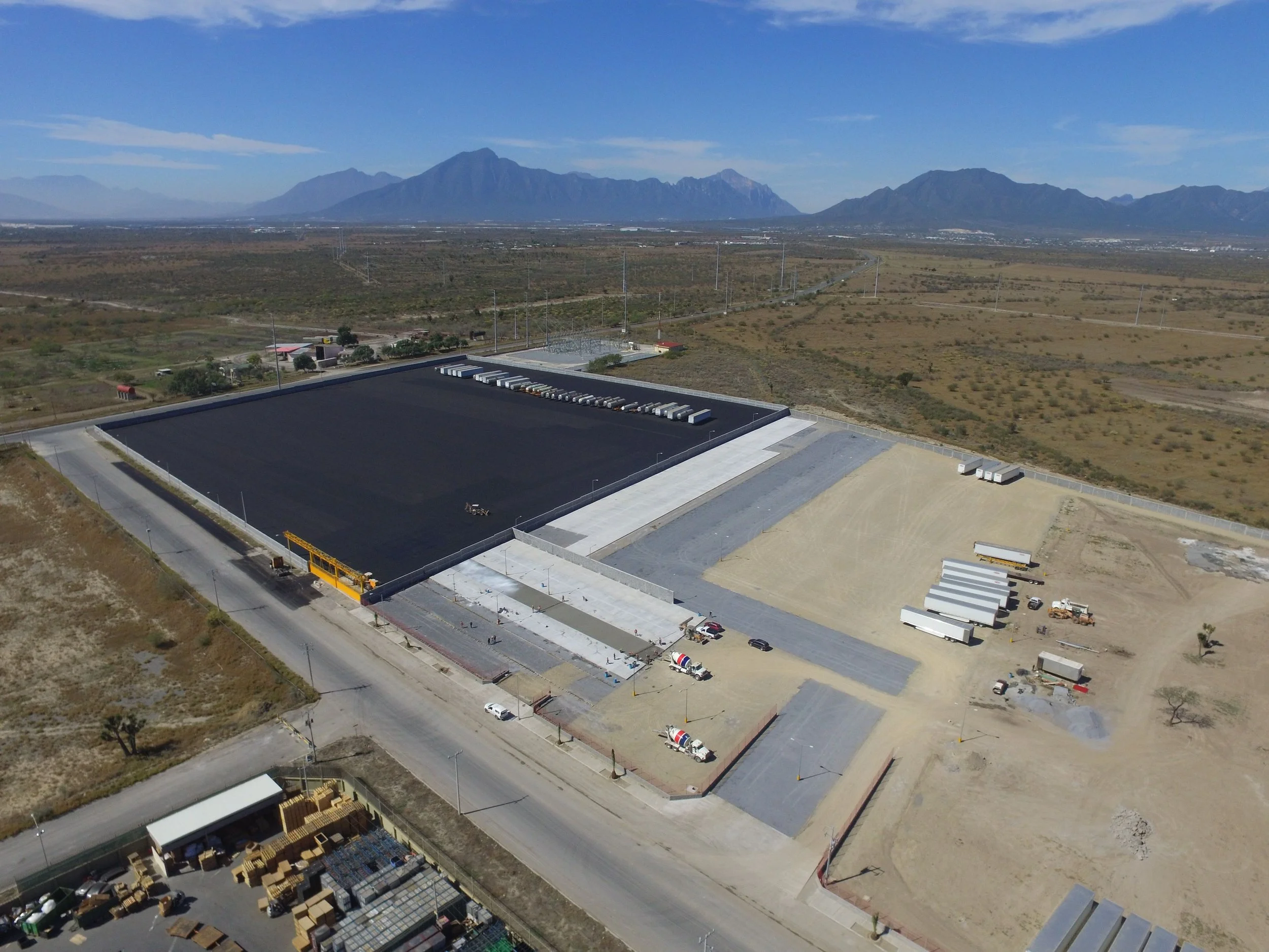 Aerial view of a construction site in a desert landscape with mountains in the background, featuring a large black asphalt area, trucks, and construction materials.