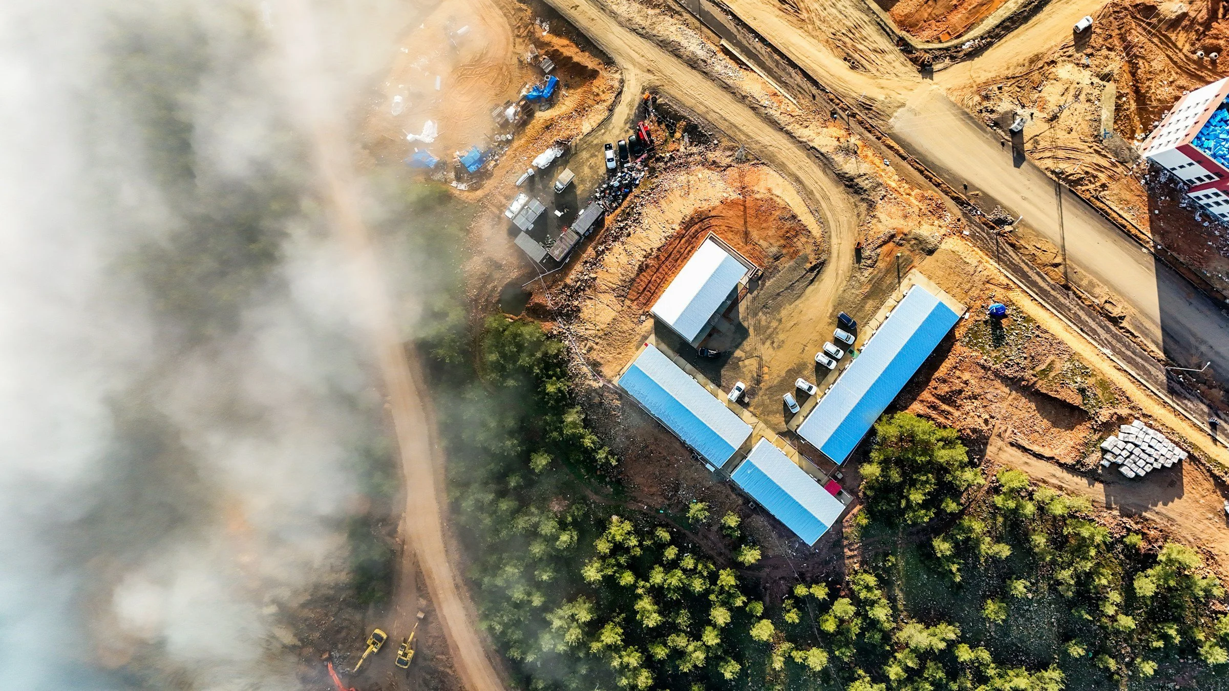 Aerial view of a construction site with dirt roads, construction vehicles, and several buildings with blue roofs, partially obscured by smoke or fog.