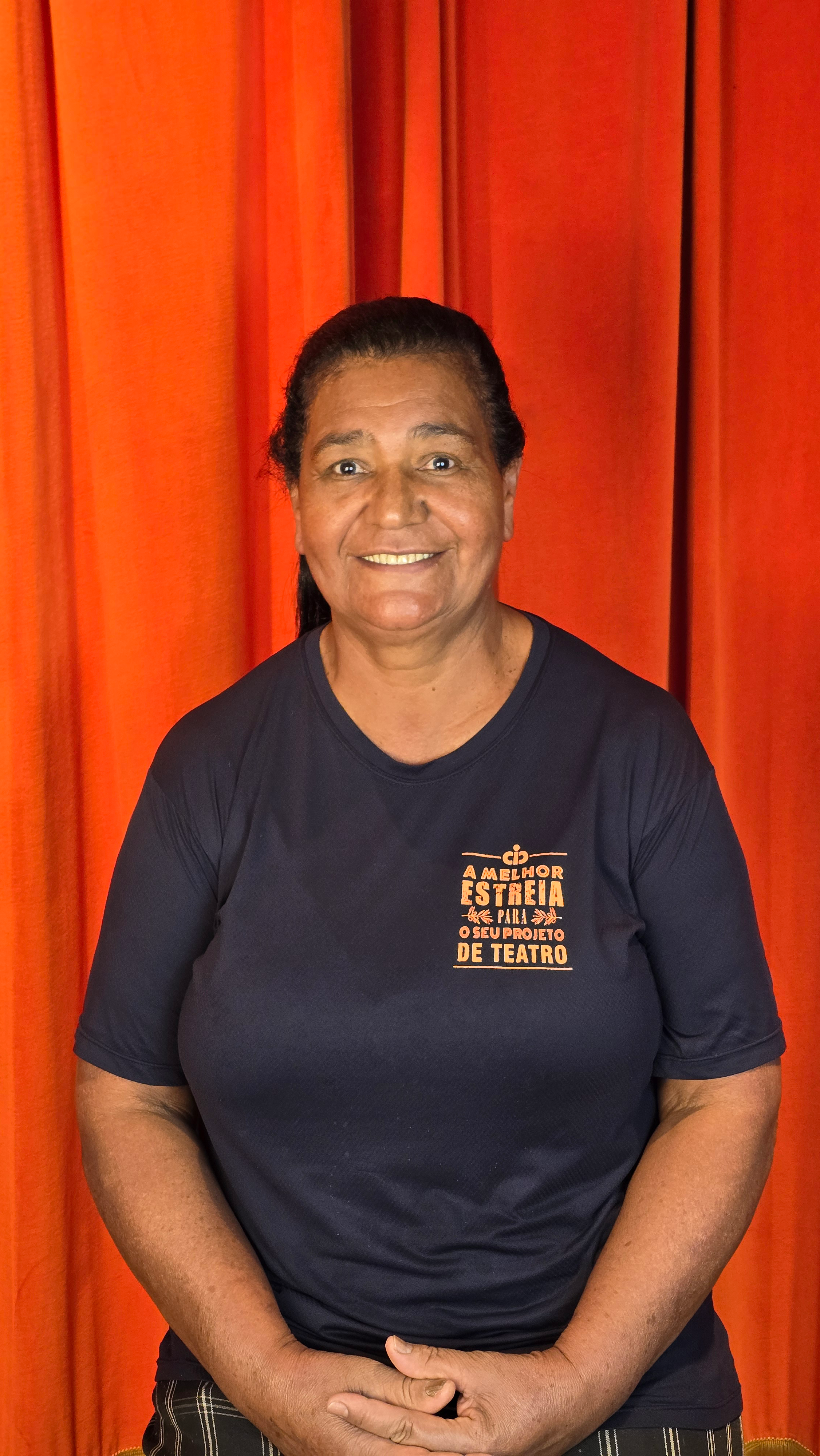 Mulher com cabelo escuro, sorriso, usando camiseta preta com uma frase em português, em pé em frente a cortinas vermelhas.