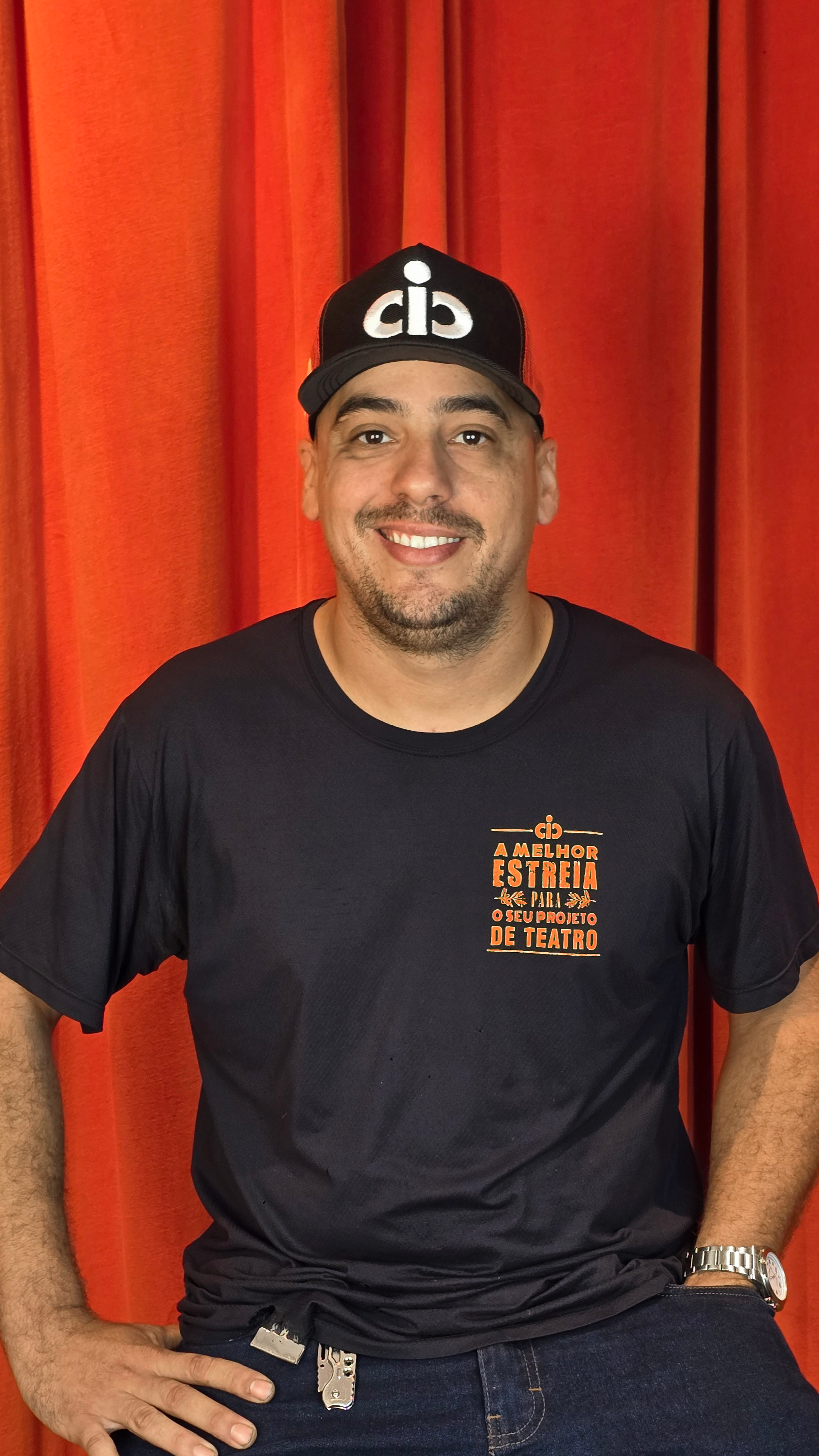 Homem sorridente usando boné preto com logotipo branco, camiseta preta com mensagem em laranja e fundo de cortina vermelha.