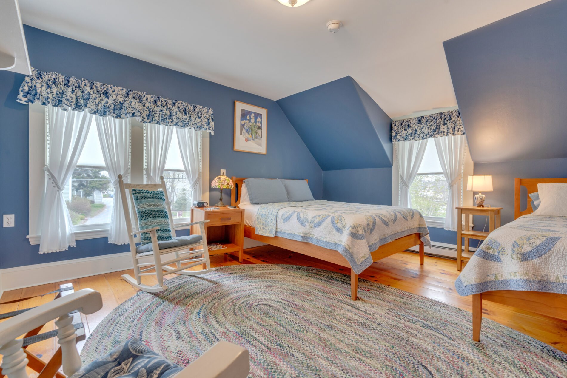 A cozy bedroom with two beds, blue walls, and large windows with white curtains and blue floral valances. There is a wooden rocking chair in the foreground, a small bedside table with a lamp, and framed artwork above the beds. The room has hardwood flooring and a colorful braided rug.