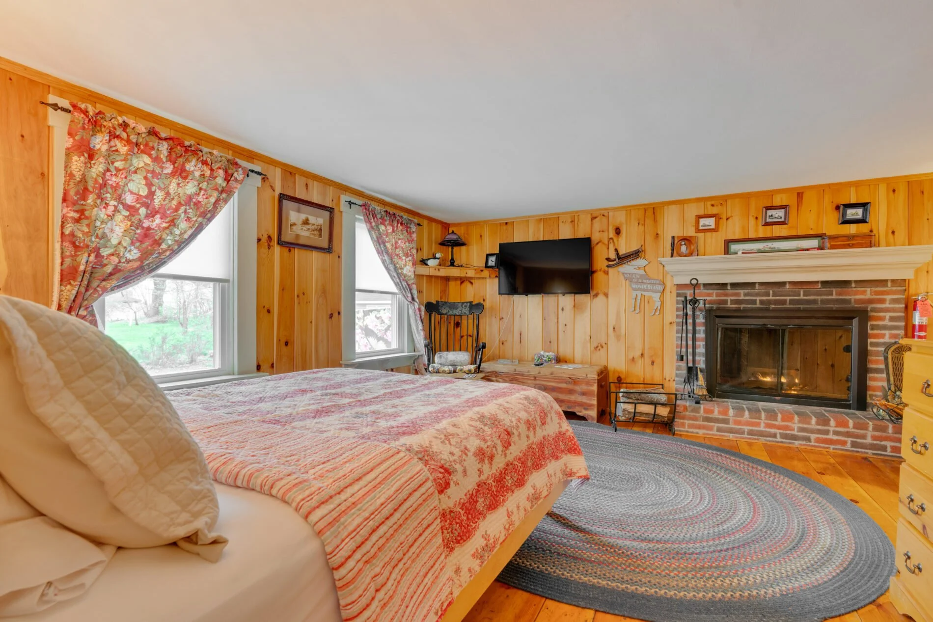 A cozy bedroom with wood-paneled walls, floral curtains, a bed with a quilt, a fireplace, a flat-screen TV, and framed pictures.