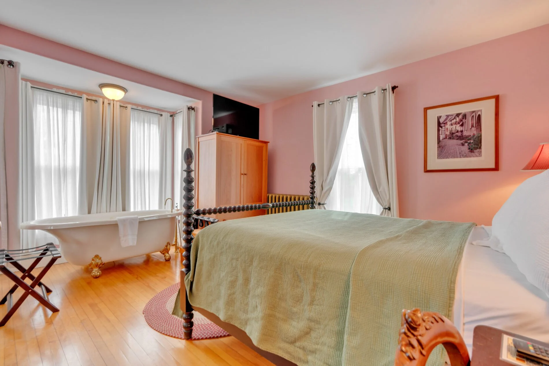 A bedroom with pink walls, a bed with a green bedspread, a wooden wardrobe, a framed picture on the wall, windows with white curtains, and a vintage-style clawfoot bathtub near the windows.