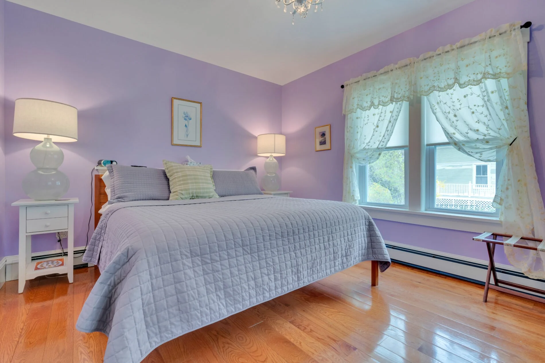 A cozy bedroom with light purple walls, a white bed with gray bedding and pillows, two white bedside tables with lamps, framed artwork on the walls, a window with lace curtains, and hardwood floors.