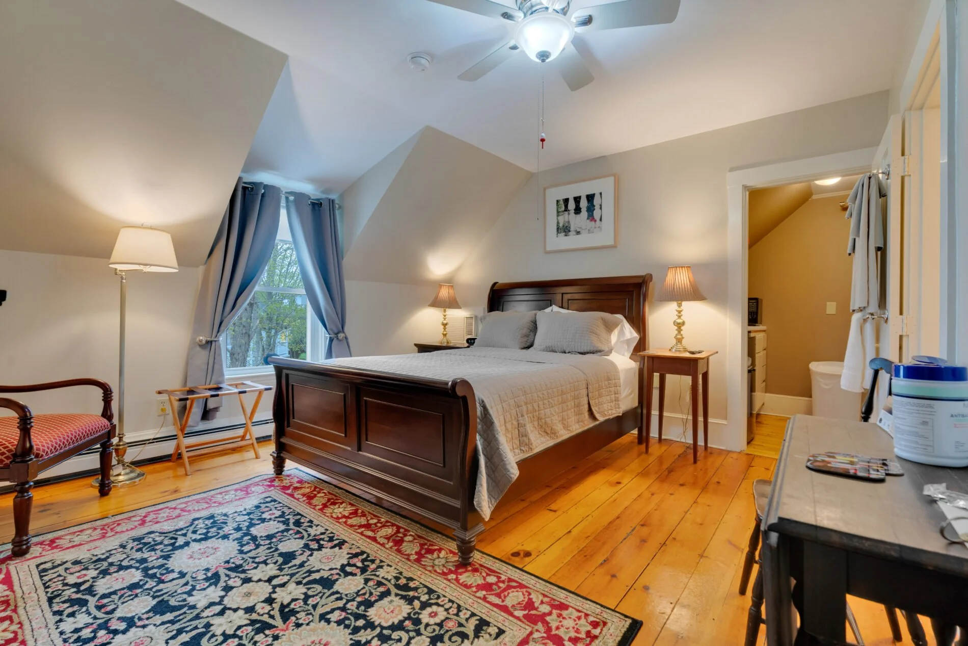 A cozy bedroom with a wooden bed frame, beige bedspread, and pillows. There are two table lamps on nightstands, a window with blue curtains, and a colorful area rug on hardwood floors. A ceiling fan with a light is on the ceiling, and an art piece hangs above the bed. To the right, there is an open door leading to a small bathroom with a toilet and beige walls. A chair with a patterned seat and a small table with personal items are also visible.