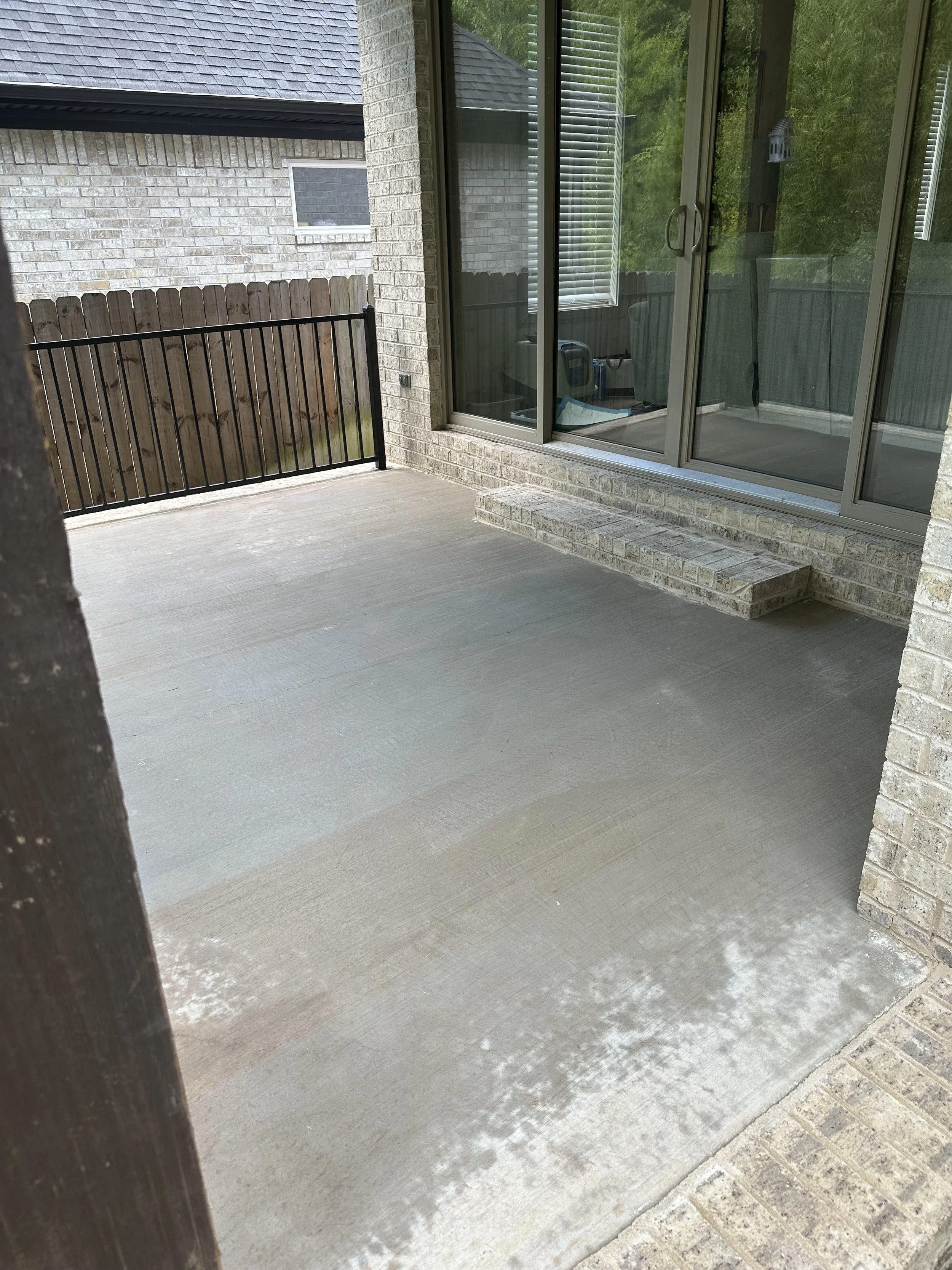 A small patio area outside a house with a sliding glass door, brick exterior walls, and a concrete floor. There is a black metal railing along the left side and a stair step leading up to the door. Some interior objects are visible through the glass door.