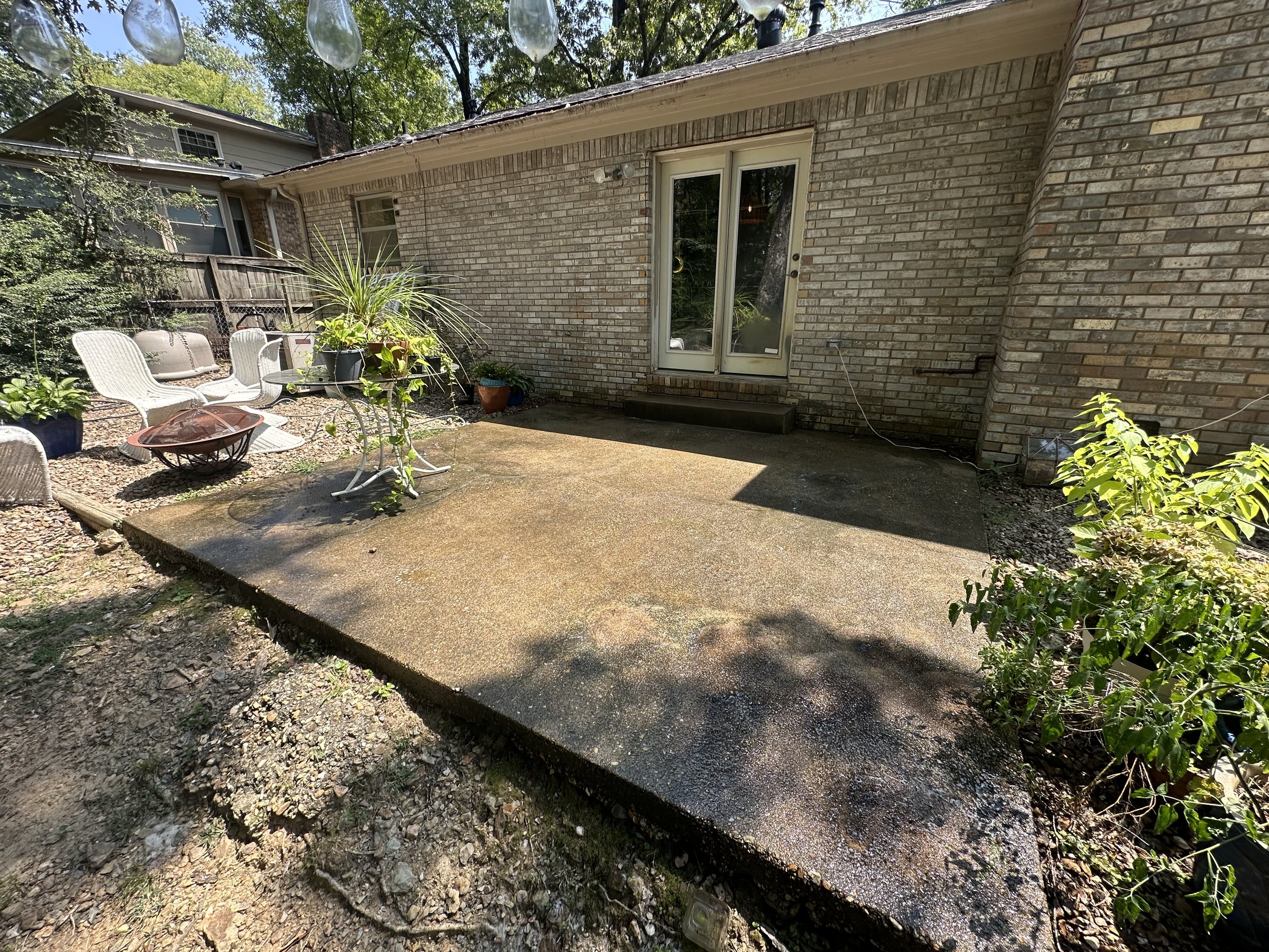 Backyard patio concrete slab before it has been pressure washed, white patio chairs, potted plants, and a brick house with a glass door, surrounded by trees and greenery.