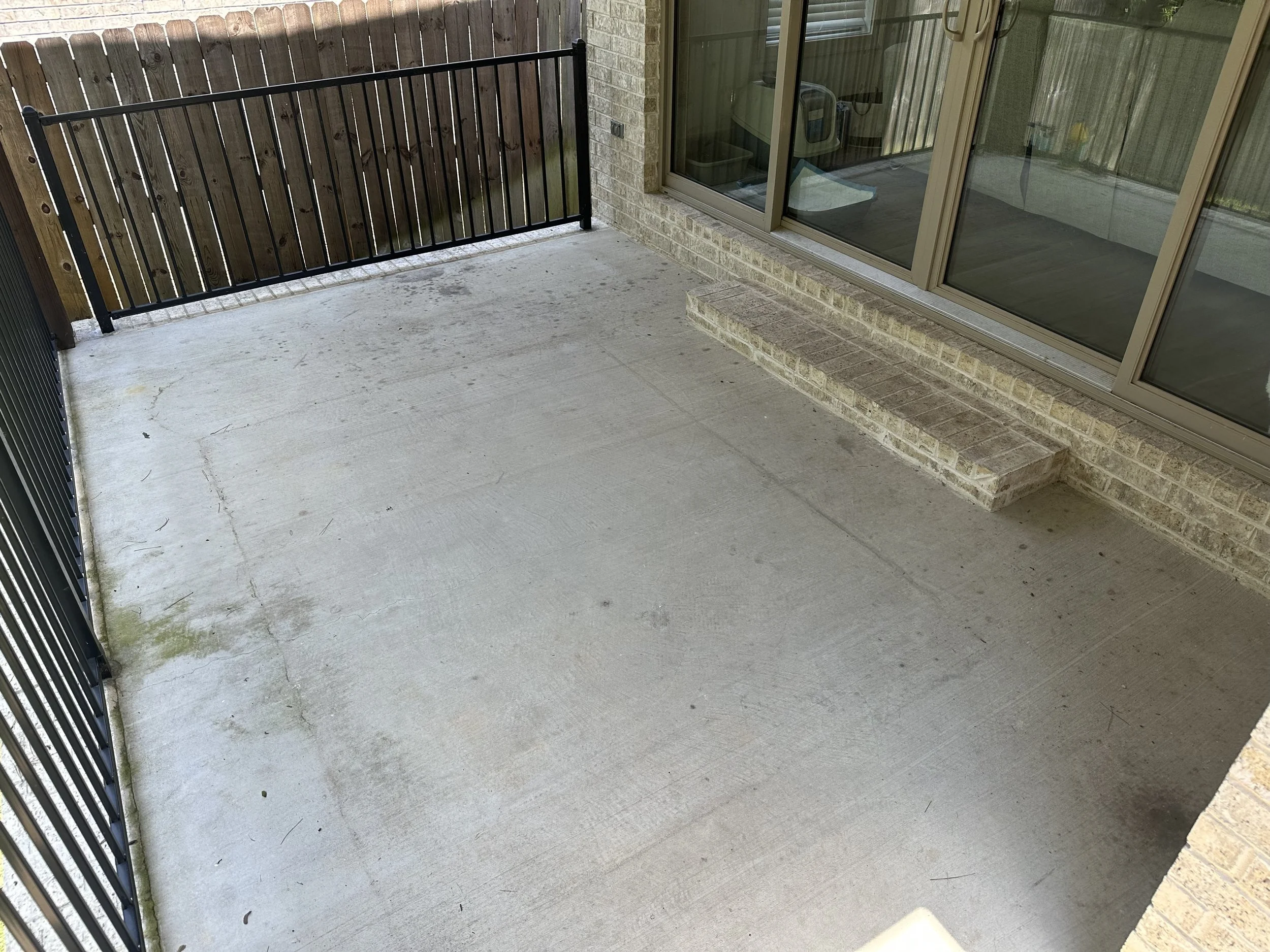 View of a small outdoor patio area with concrete flooring before it has been pressure washed, a wooden fence, a sliding glass door, and a brick wall. There are some steps leading up to the door.