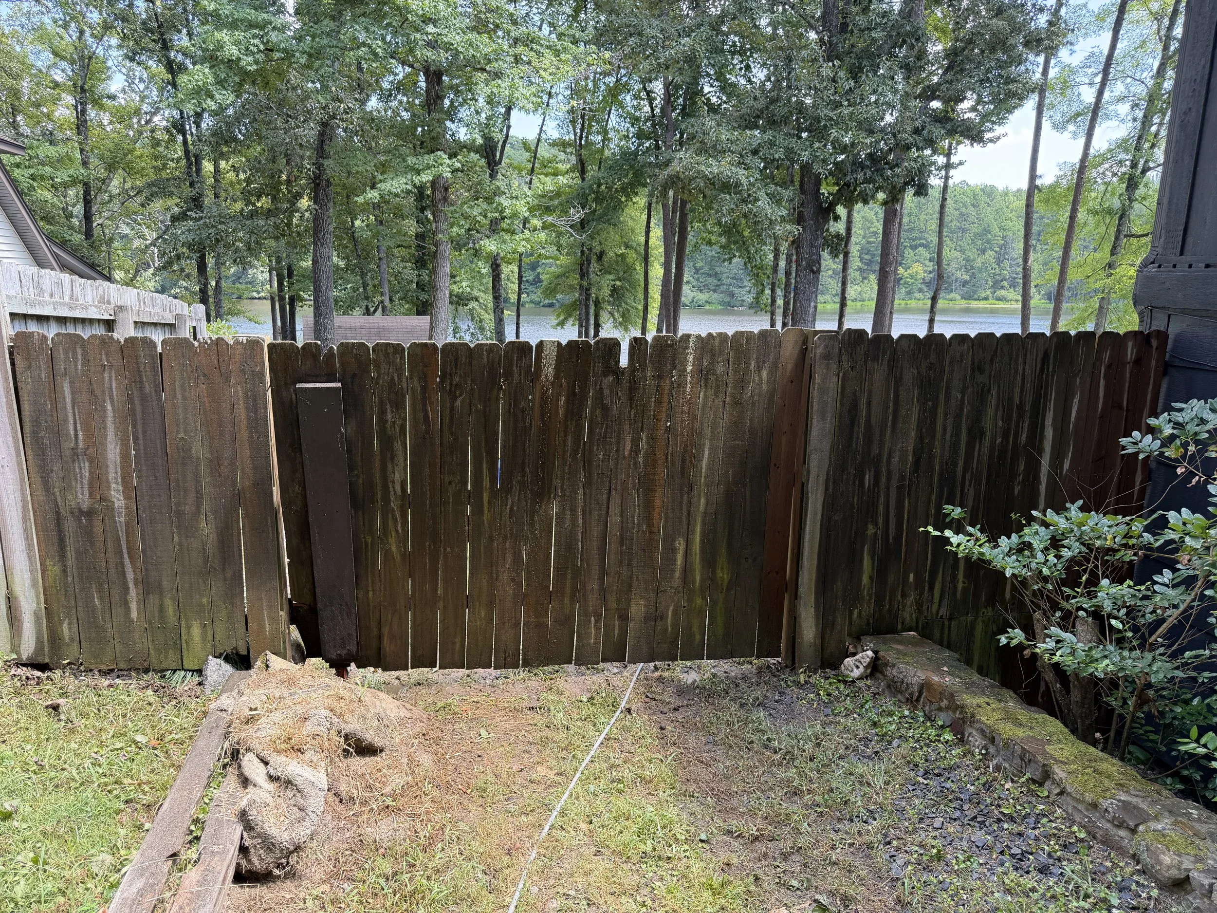 View of a wooded backyard with a wooden fence before it has been pressure washed, trees, and a lake in the background, partially visible through the trees.