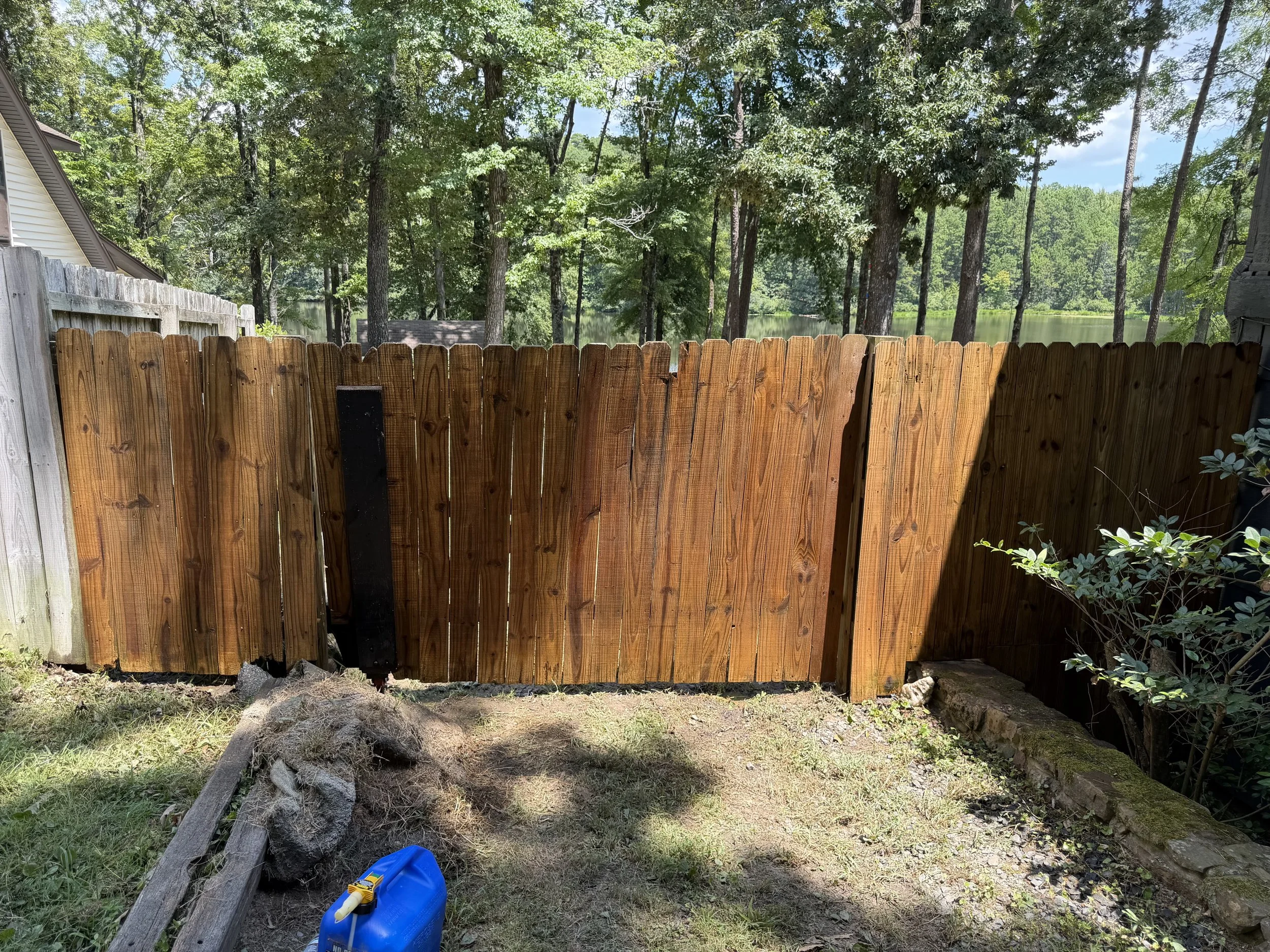 A backyard with a wooden fence that has been freshly pressure washed, trees, and a lake in the background. There is a blue watering can and some wood planks on the ground.