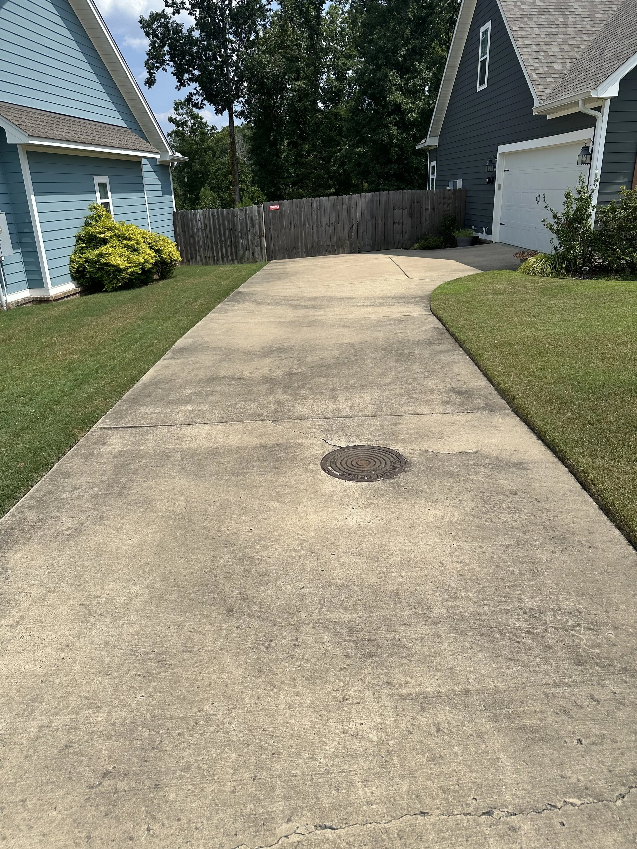 Concrete driveway before it has been pressure washed, heading to two houses with lawns and bushes, wooden fence, and trees in the background.