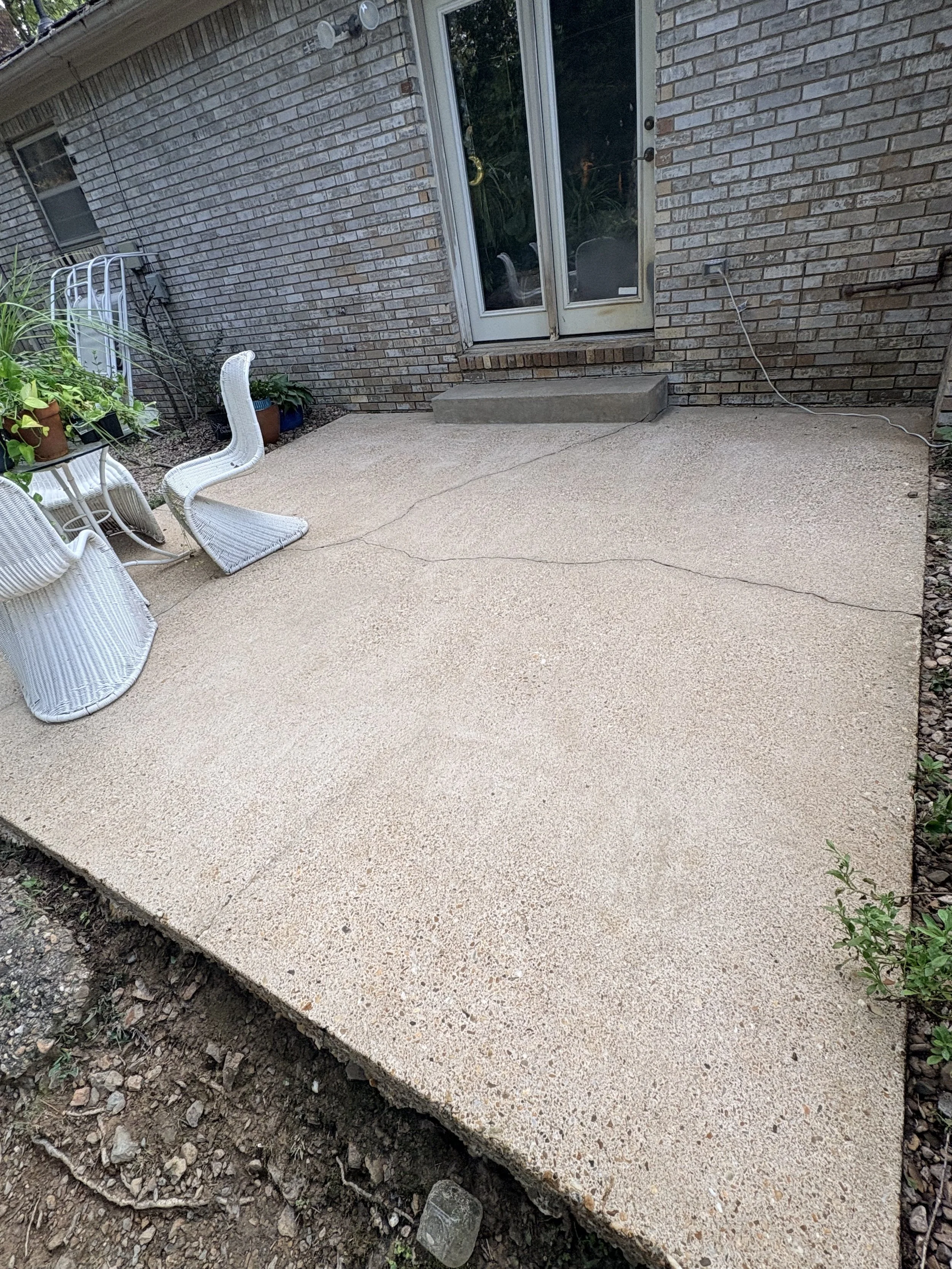 A backyard patio freshly pressure washed, adjacent to a brick house with glass doors. There are white outdoor chairs and potted plants on the patio, with a garden border along the edge.