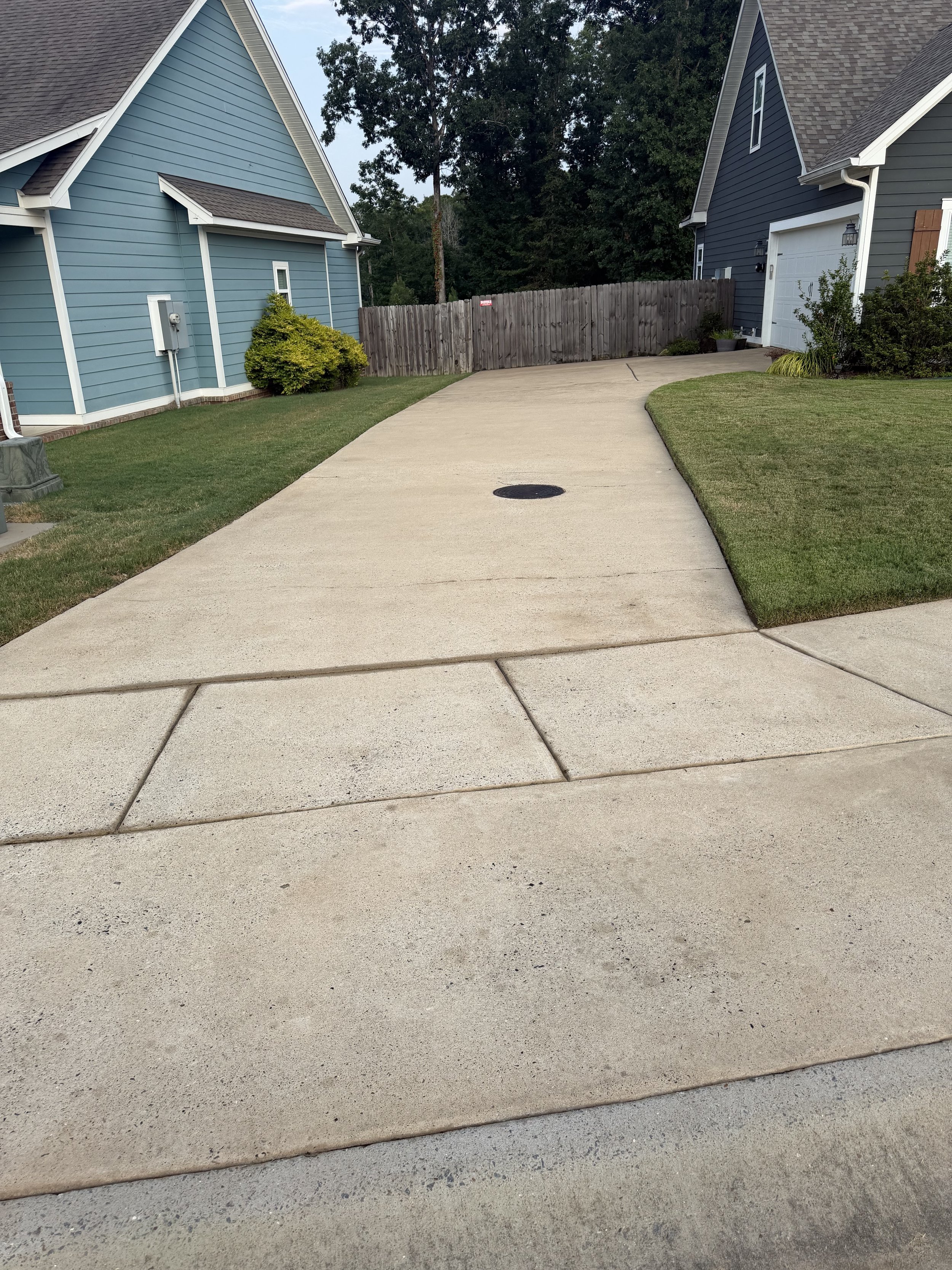 Residential driveway freshly pressure washed with grass on the sides, two blue houses with white trim, some bushes, a wooden fence at the end of the driveway, and trees in the background.