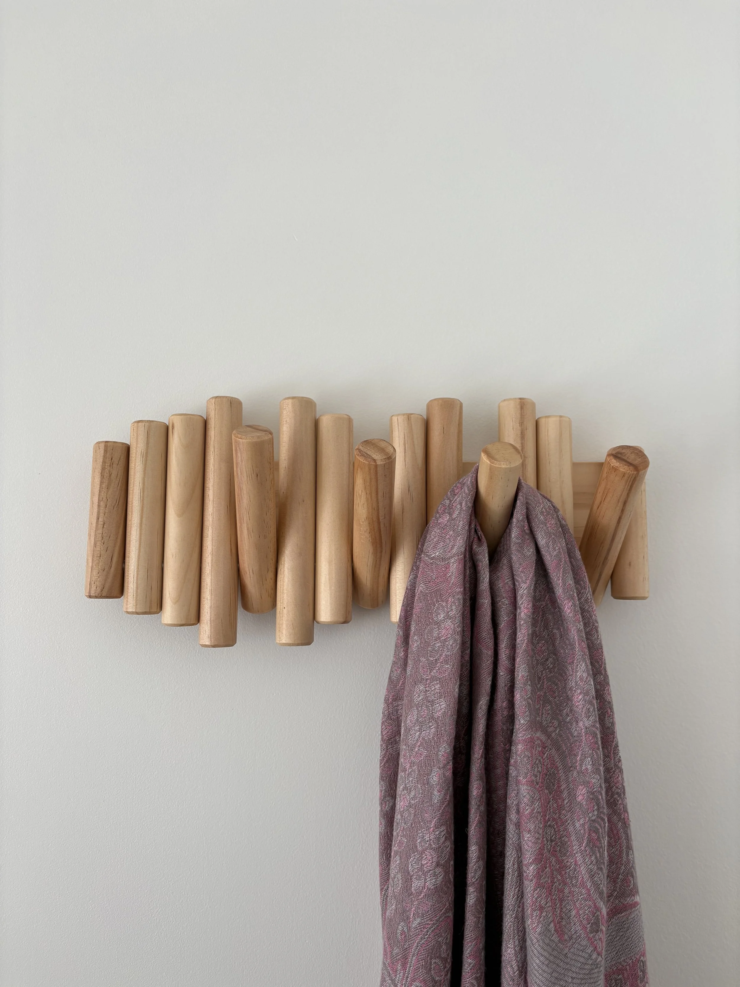 Wooden coat rack with several vertical sticks and a pink patterned scarf hanging from it, against a plain white wall.