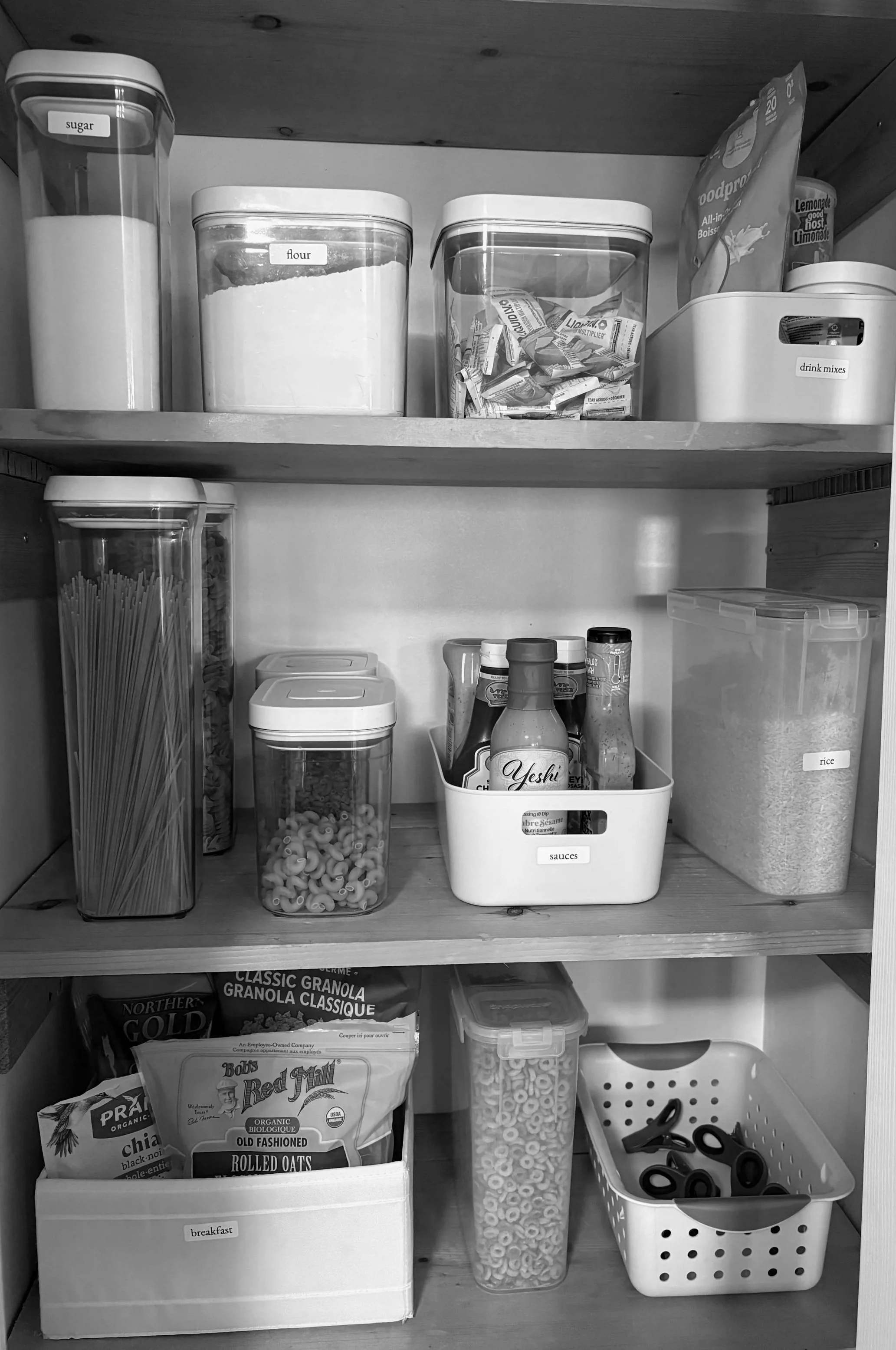 Organized kitchen pantry with labelled containers holding sugar, flour, dry drink mixes, rice, pasta, cereal, and sauces.