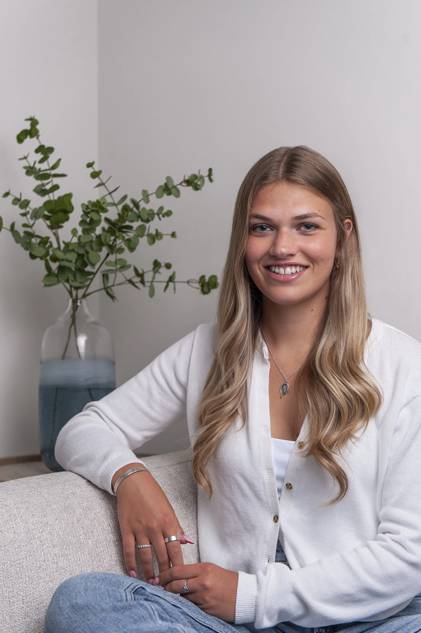 A young woman with long blonde hair, wearing a white cardigan, sitting on a beige couch with an arm resting on it, smiling at the camera, with a vase of leafy green plants in the background.