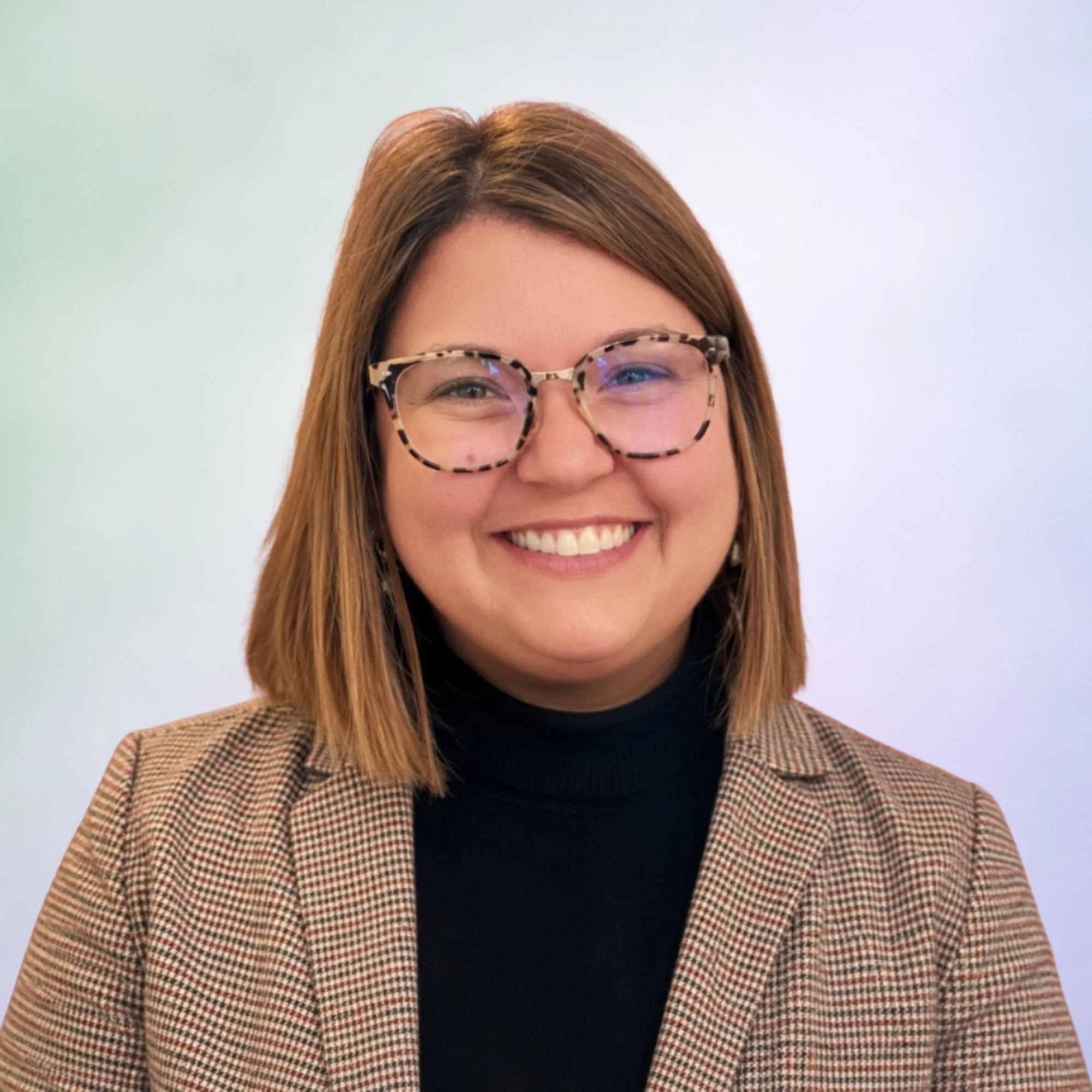 Close-up of a smiling woman with light brown hair wearing leopard print glasses and a checked blazer, against a light background.