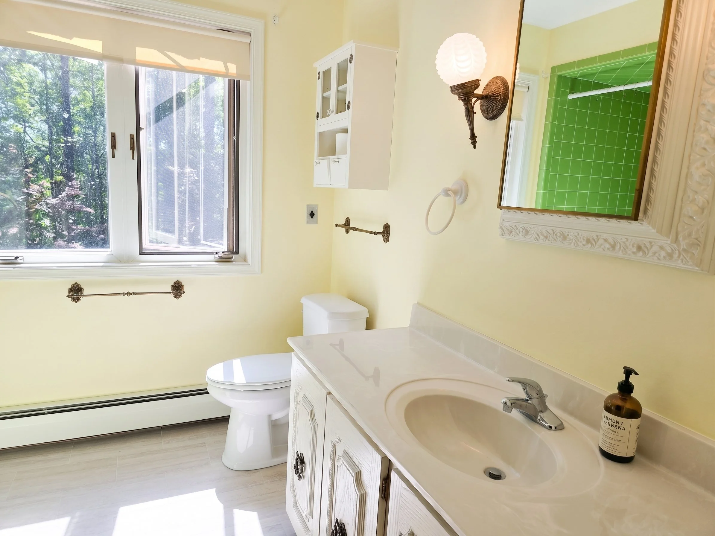 Bright, clean sober living residence bathroom with white vanity, light yellow walls, large window, and gold-framed mirror