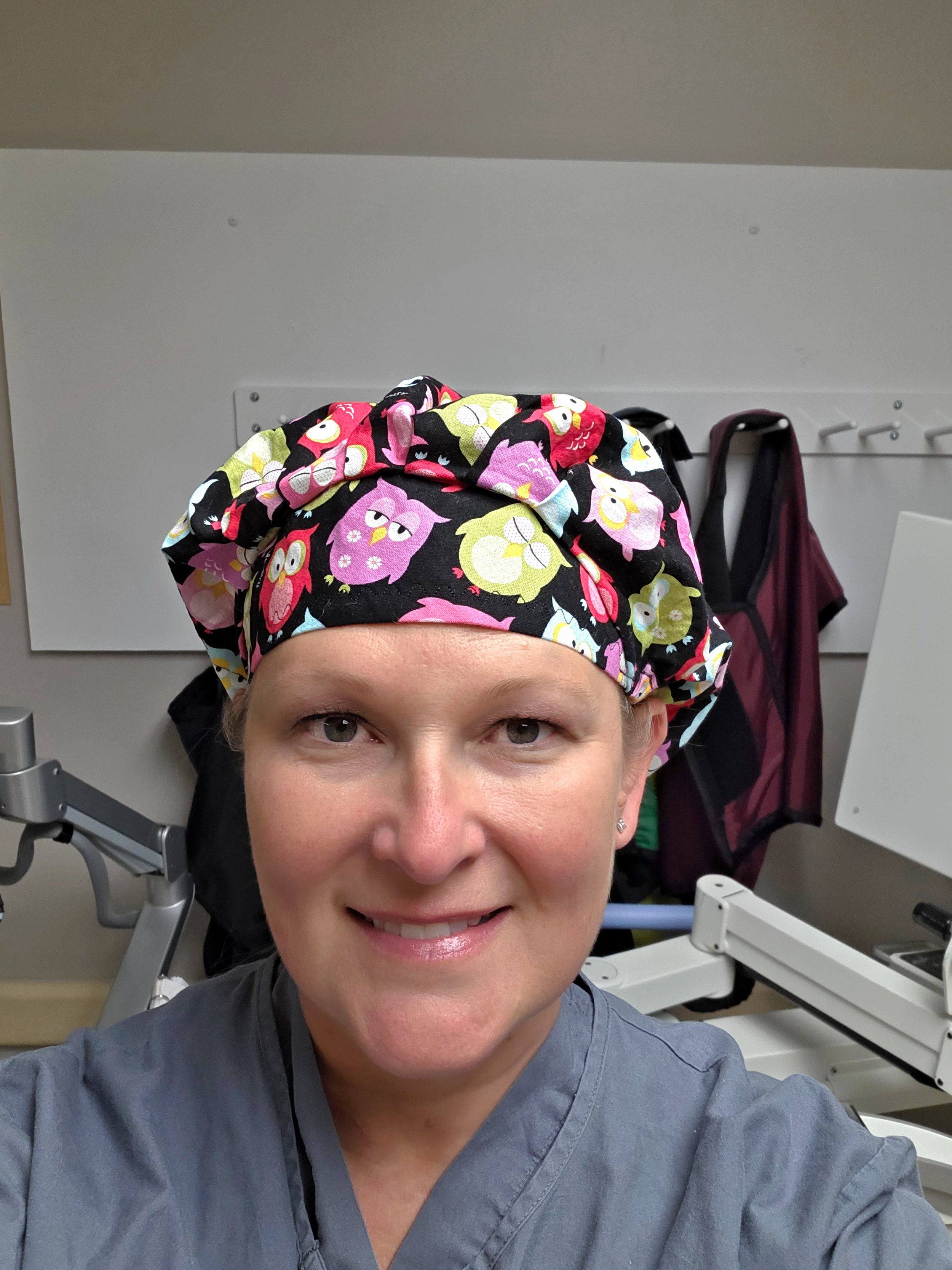 A nurse in gray scrubs taking a selfie in a hospital room with medical equipment in the background and a woman wearing a colorful owl scrunchie on her head.