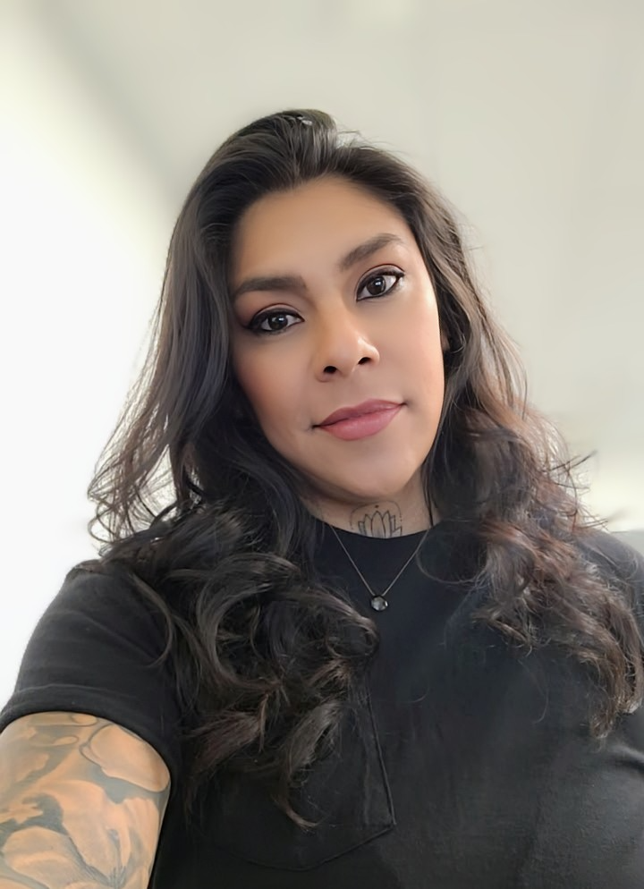 A woman with long, dark, curly hair taking a selfie indoors. She is wearing a black top, a small necklace with a pendant, and has a tattoo on her arm and neck.
