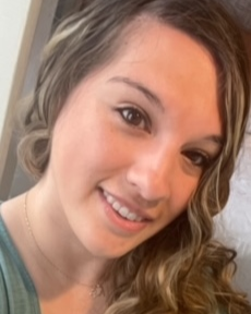 A woman smiling with long curly hair, wearing a light-colored top.