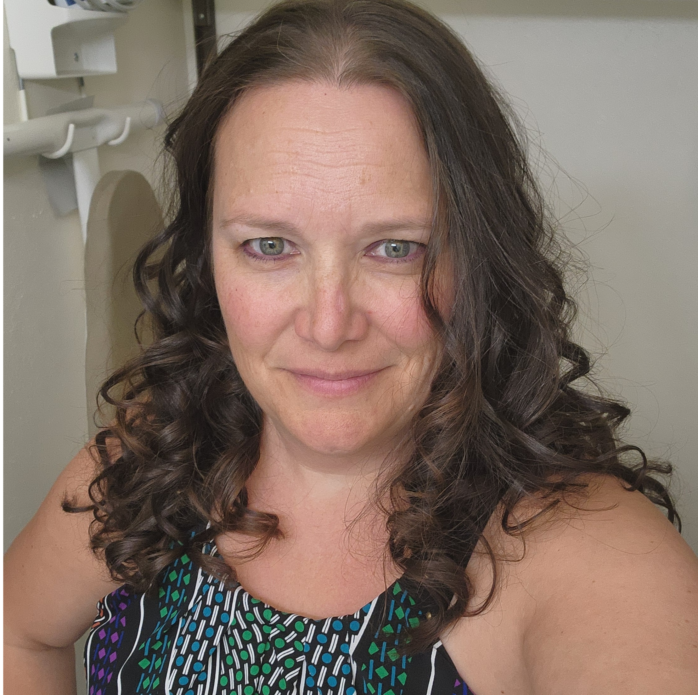 A woman with long, curly brown hair and light skin is smiling slightly and looking into the camera. She is wearing a colorful, patterned top. The background includes part of a white wall and a medical or dental device.