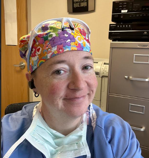 Person wearing colorful scrubs and a matching cap, smiling in an office or medical room setting.