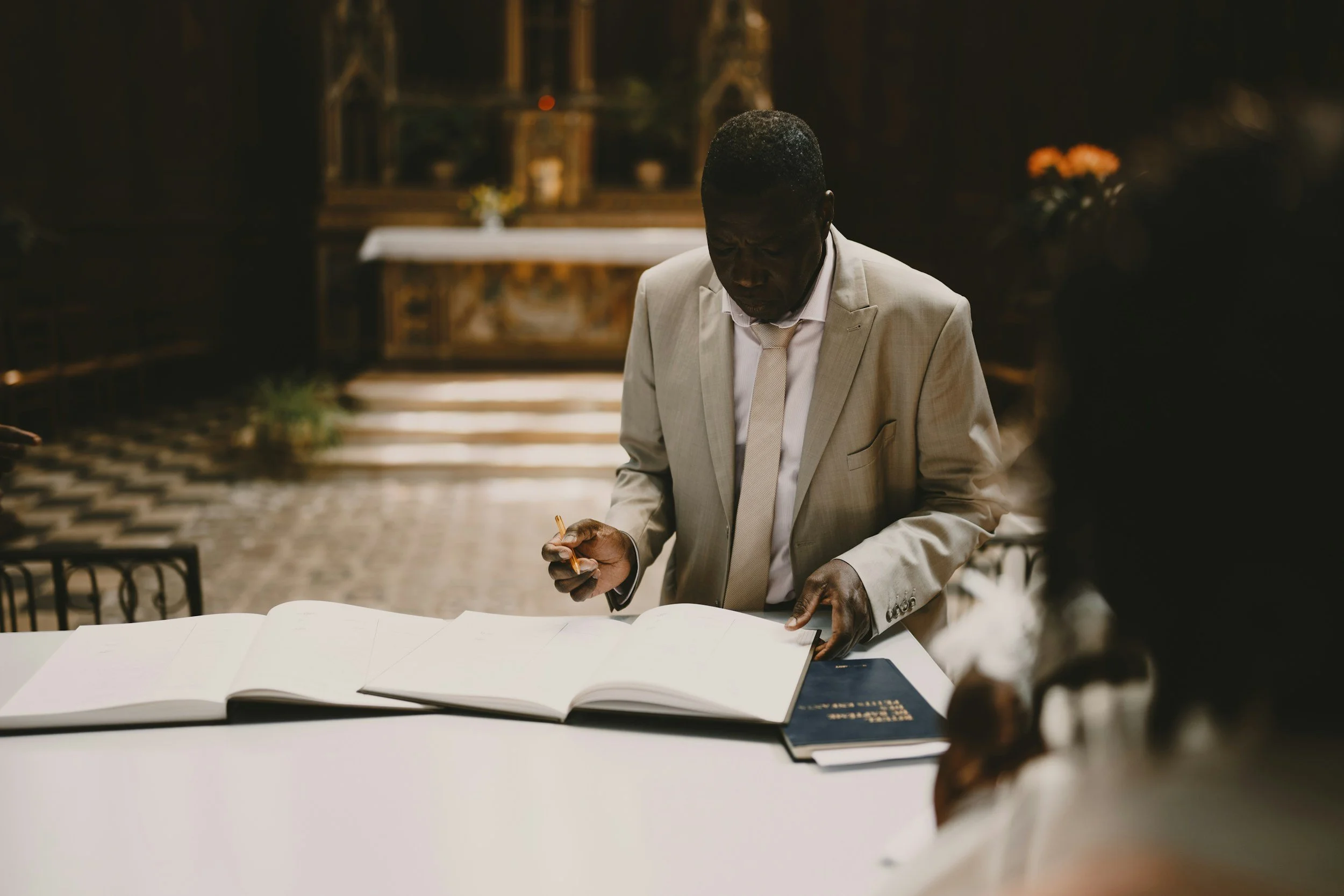 A man in a light-colored suit signing a document at a table in a dimly lit room, with a church altar in the background.