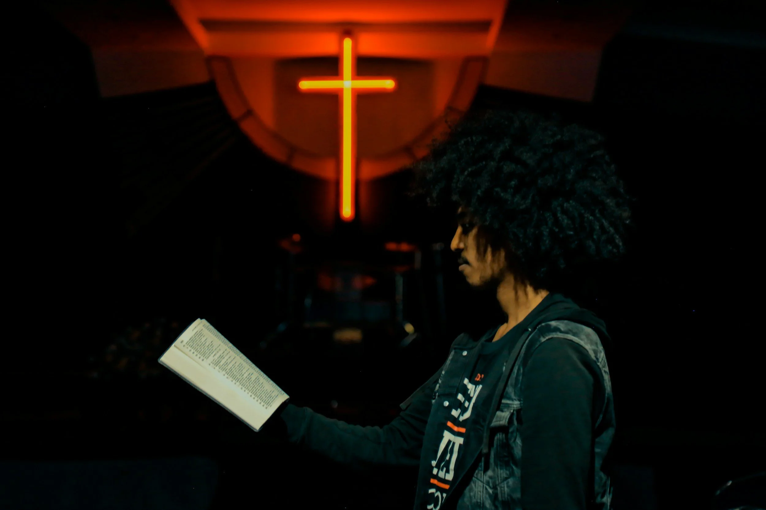 A person with curly hair is reading a book or booklet in a dark room illuminated by the red glow of a neon cross on the wall behind them.