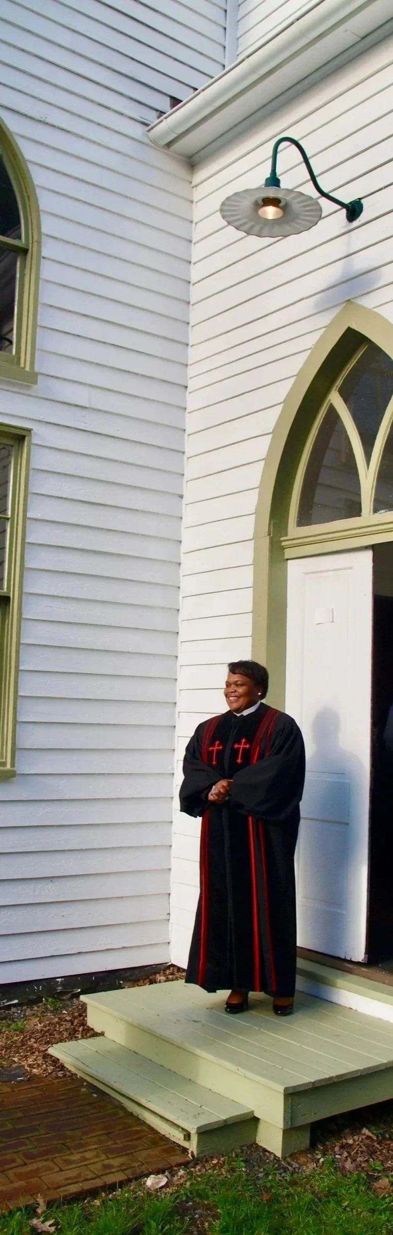 A woman dressed in a black choir robe with red crosses stands on a small green porch outside a white church. She is smiling and clasping her hands in front of her.