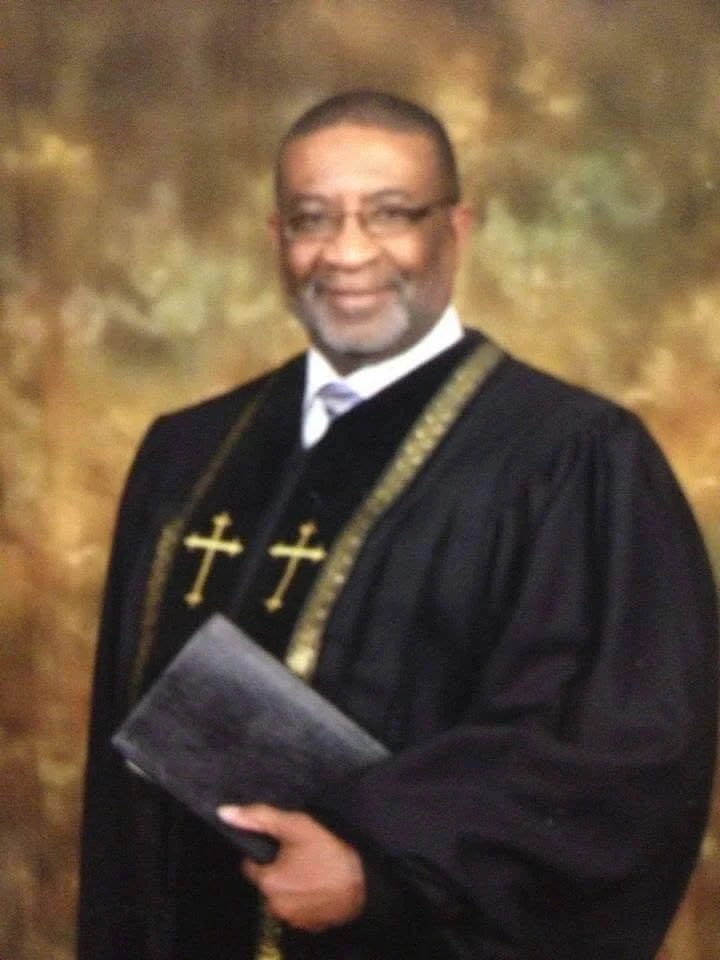 A man in a black religious robe with gold cross necklaces, holding a book, standing in front of a gold and brown background.