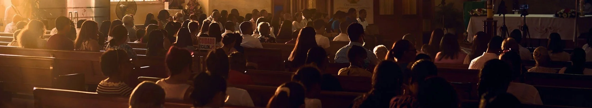 A congregation of people seated in pews inside a church, sitting quietly during a service with warm sunlight streaming through windows.
