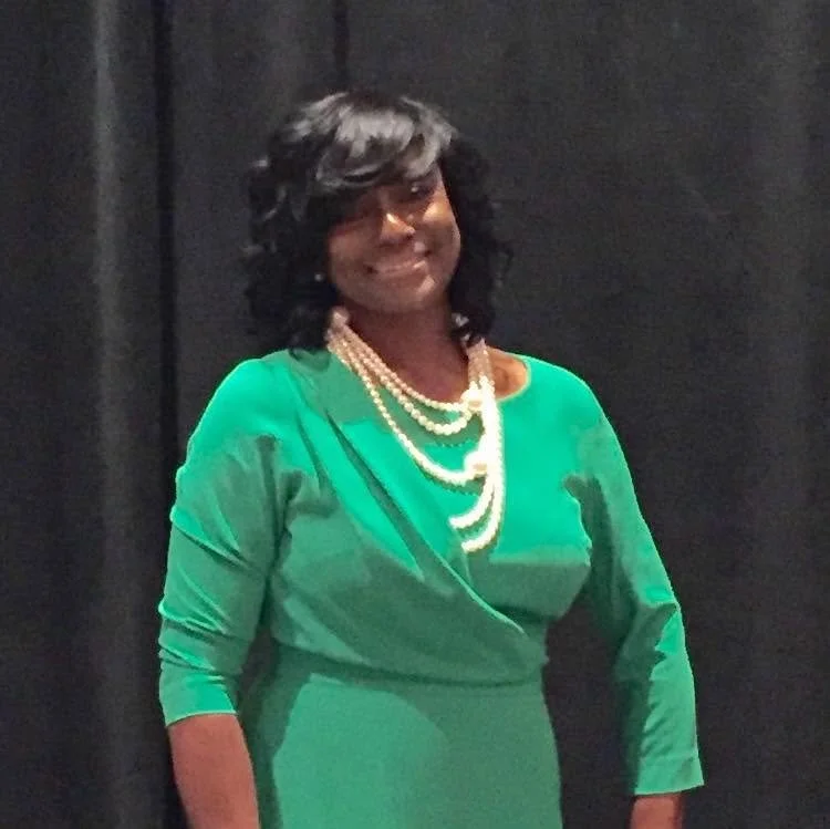 A woman with black, wavy hair, wearing glasses, a green dress, and a pearl necklace, smiling at the camera against a dark background.