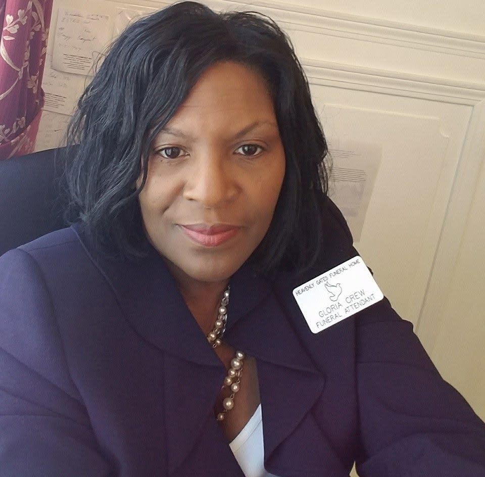 A woman with short black wavy hair and medium-brown skin, dressed in a navy blazer and pearl necklace, sitting and looking at the camera in an office setting. She has a name tag that reads 'Gloria Crew, Funeral Attendant,' and a small penguin drawing.
