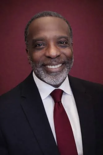 A middle-aged African American man with gray hair and beard, smiling, dressed in a dark suit, white shirt, and red tie, against a burgundy background.