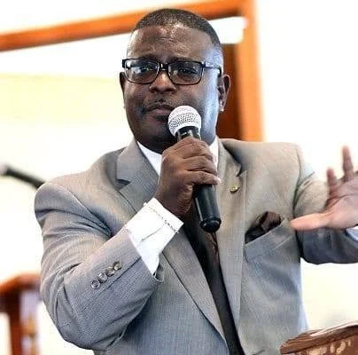 Man in gray suit and glasses speaking into a microphone during a presentation or speech.