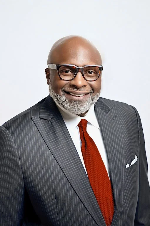 A smiling African American man in a dark pinstripe suit, white shirt, red tie, glasses, and a white pocket square, standing against a light background.