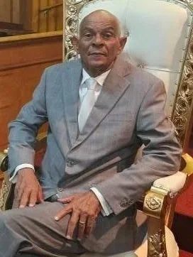 An elderly man in a light gray suit sitting on an ornate chair with gold accents in a formal setting.