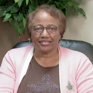 An elderly woman wearing glasses, a black shirt with a silver star, and a light pink cardigan, sitting indoors with green foliage in the background.