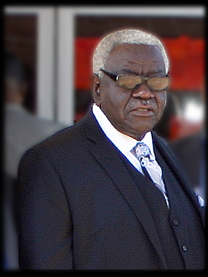 An elderly man with gray hair and glasses wearing a dark suit and tie.