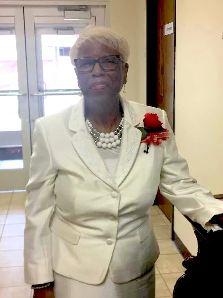 An elderly African American woman with short white hair, wearing glasses, a white suit, a pearl necklace, and a red corsage, standing indoors near a door.