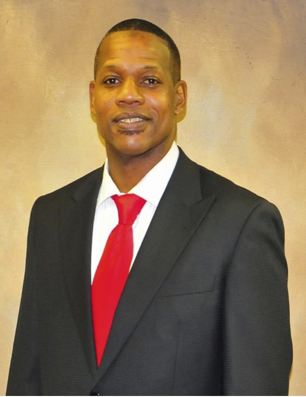 A man in a black suit with a white shirt and red tie, standing against a beige background.