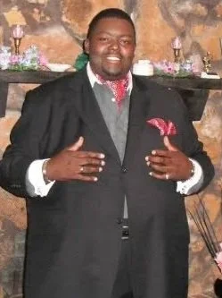 A man dressed in a formal suit smiling and making a gesture with both hands in front of a decorated background.