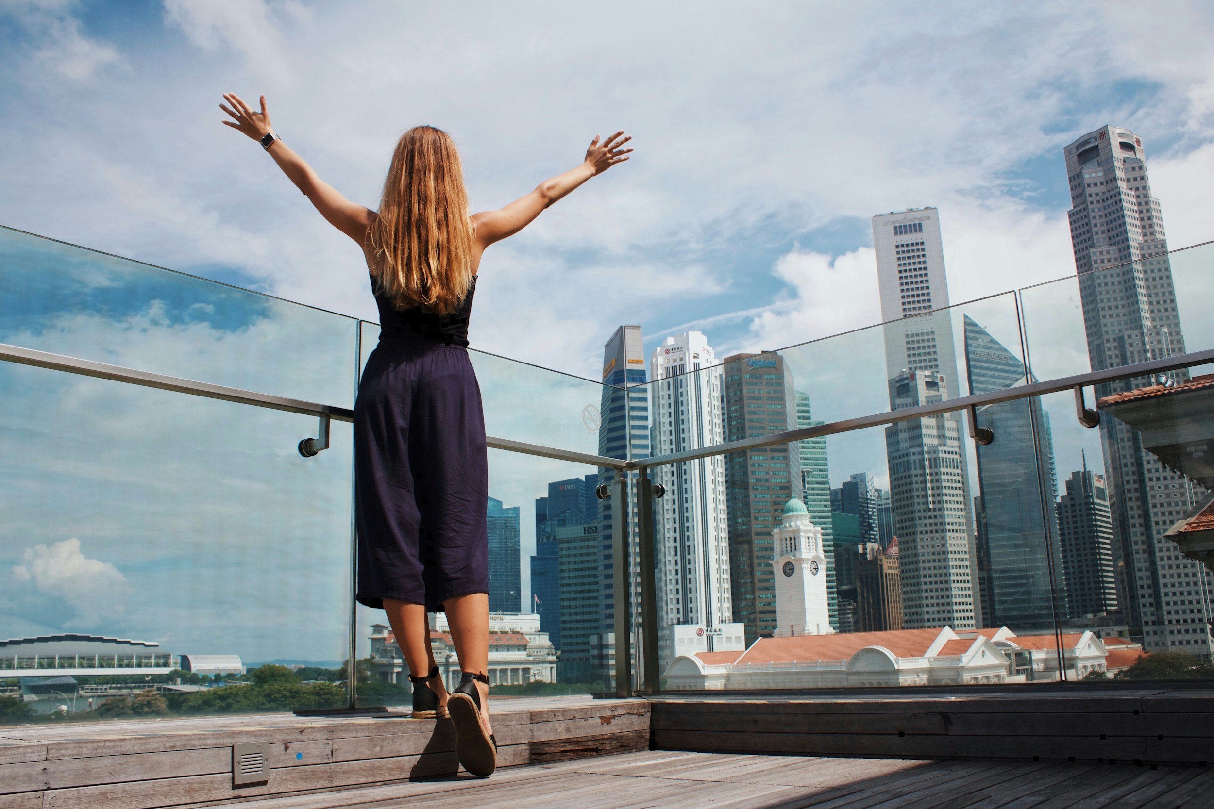 A woman with long red hair wearing a black top and purple pants stands on a rooftop terrace with glass railings, overlooking a city skyline with tall buildings under a partly cloudy sky. She has her arms raised and facing away from the camera.