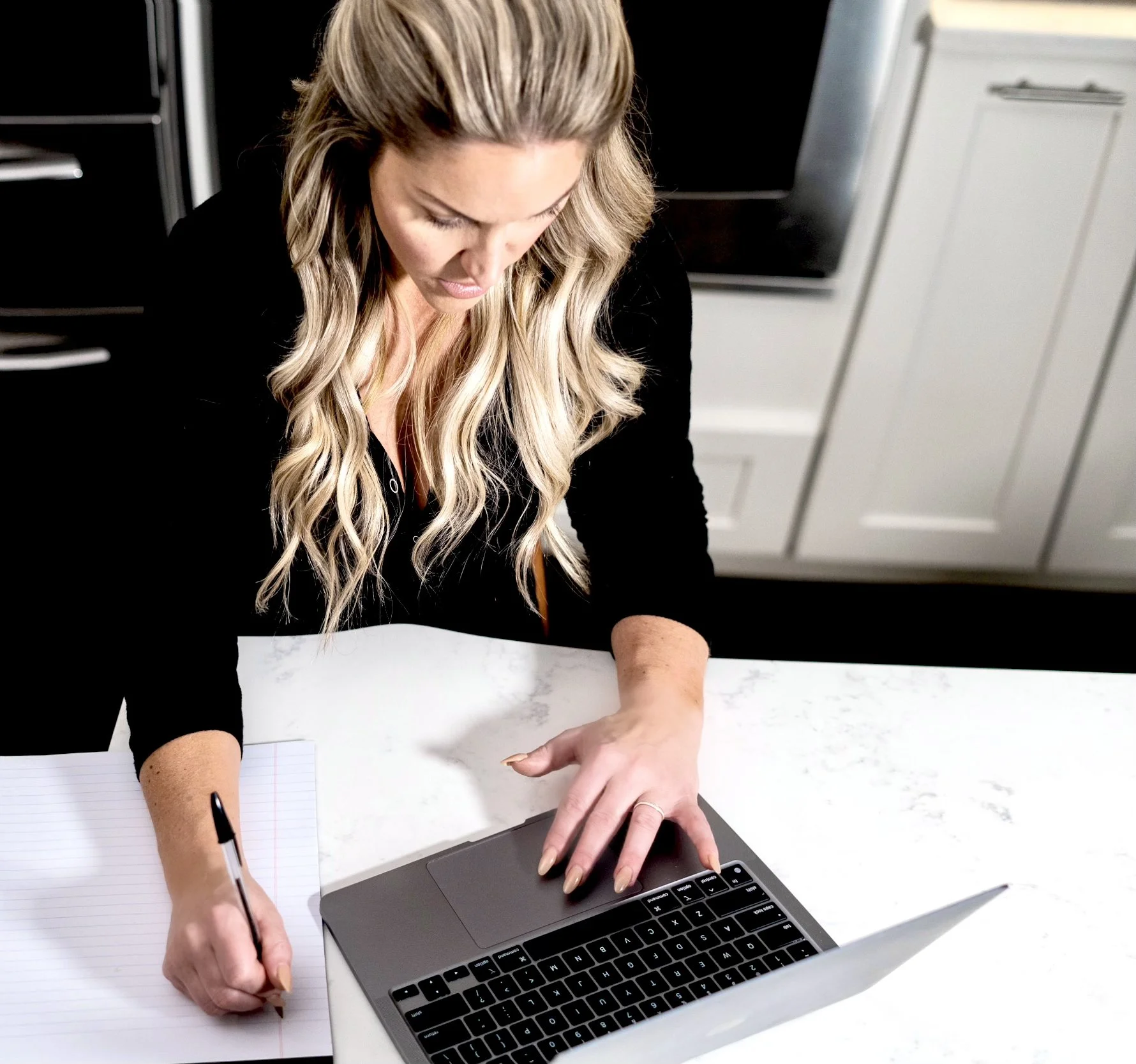 A woman with long wavy blonde hair working on a laptop at a white marble desk, writing in a notebook with a black pen.
