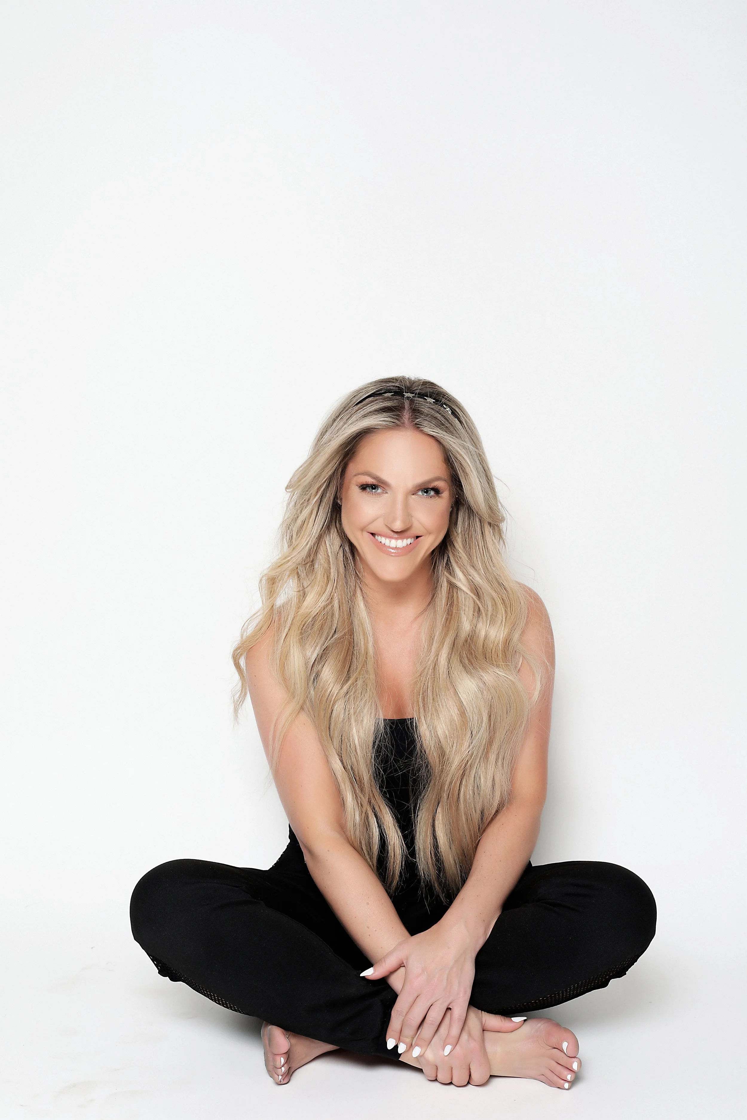 A smiling woman with long, wavy blonde hair, sitting cross-legged against a plain white background, wearing a black sleeveless top and black pants.